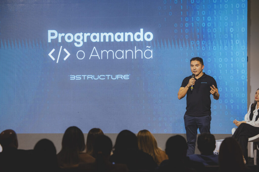 Homem branco, camisa preto e calça jeans, está com o microfone em mãos. Atrás dele, um painel de apresentação, com a frase "Programando o Amanhã". Ele faz a apresentação do projeto no Talk Gestão de Valor