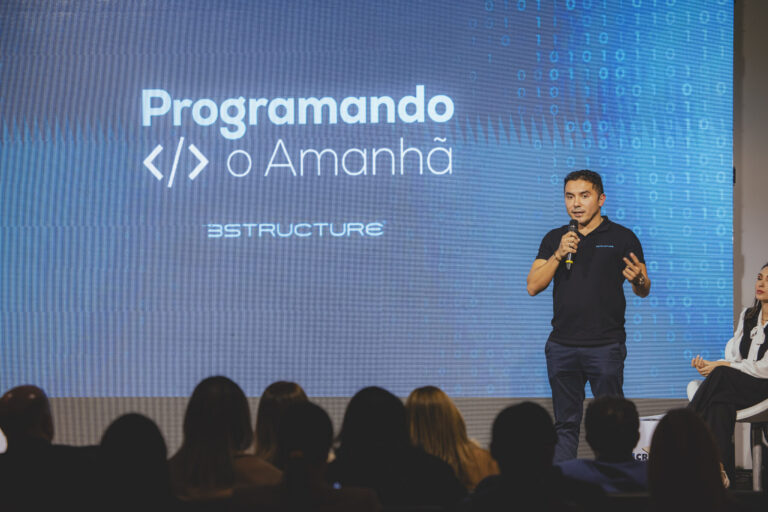Homem branco, camisa preto e calça jeans, está com o microfone em mãos. Atrás dele, um painel de apresentação, com a frase "Programando o Amanhã". Ele faz a apresentação do projeto no Talk Gestão de Valor