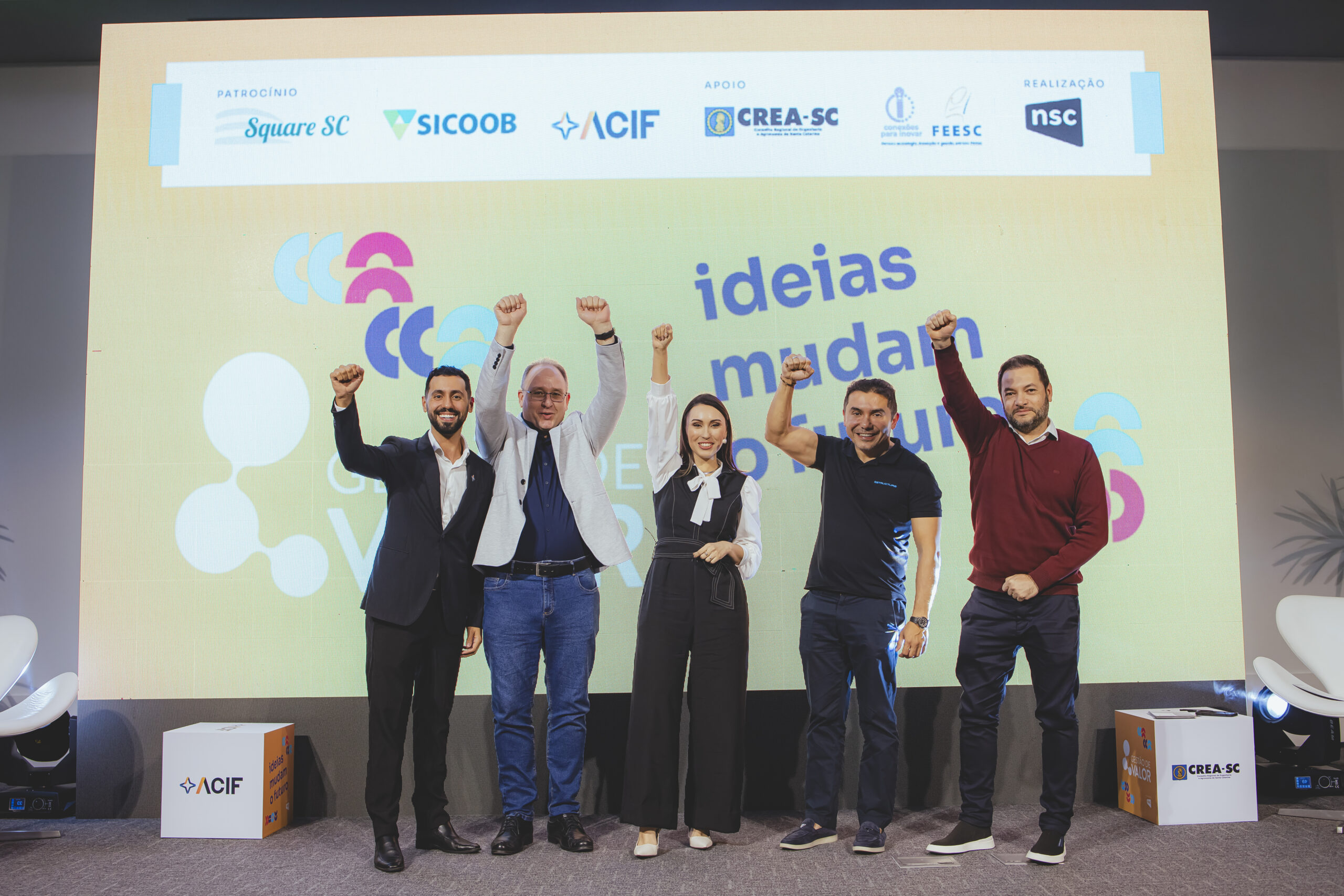 Em cima de um palco, em frente a um painel, estão cinco pessoas: quatro homens e uma mulher. Da esquerda para a direita: Homem branco, cabelos castanhos curtos, barba, sorrindo. Veste suéter vermelho, jeans e tênis. Braço esquerdo estendido para baixo, braço direito levantado, mãos fechadas. Homem branco, cabelos pretos curtos, camisa preta da Estructure, jeans e tênis. Sorrindo, braço direito levantado, mão esquerda estendida para baixo. Mulher branca, cabelos longos, camisa branca, macacão jeans escuro e sapatos brancos. Sorrindo, com microfone, braço direito levantado, mãos fechadas. Homem branco, cabelos loiros curtos, óculos, sorrindo. Veste blazer branco, camisa azul, jeans e sapatos pretos. Braços levantados, mãos fechadas. Homem branco, cabelos pretos curtos, sorrindo. Veste blazer e calça social escura, camisa branca. Braço direito estendido, braço esquerdo ao lado do corpo. No telão atrás deles, a frase "Ideias moldam o futuro". No canto inferior do telão, uma caixa quadrada com o logo da Acif (predominantemente azul com detalhes laranja), sobre fundo branco e laranja.