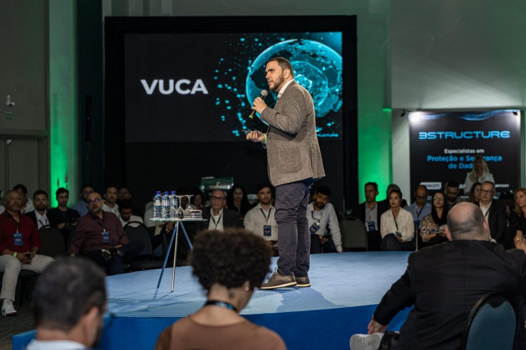 A direita do palco circular, Àquilla Amaral, veste camisa branca, blazer cinza e calças jeans.