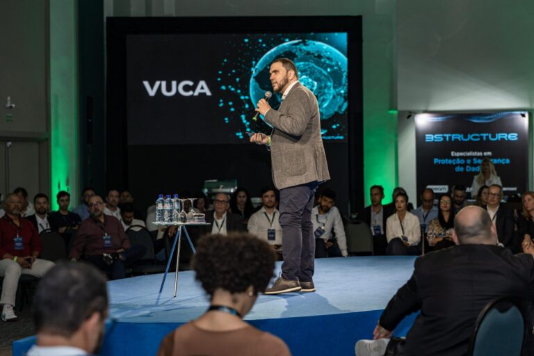 A direita do palco circular, Àquilla Amaral, veste camisa branca, blazer cinza e calças jeans.